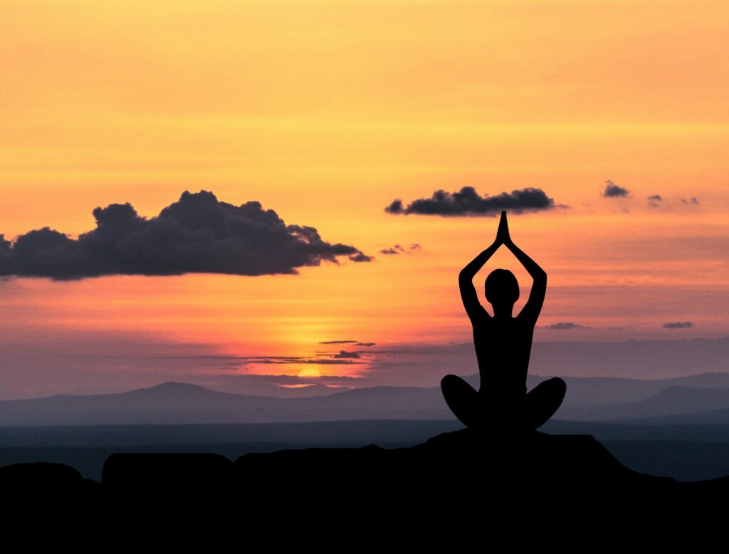 Person holds prayer above head as they sit cross-legged in front of sunset.